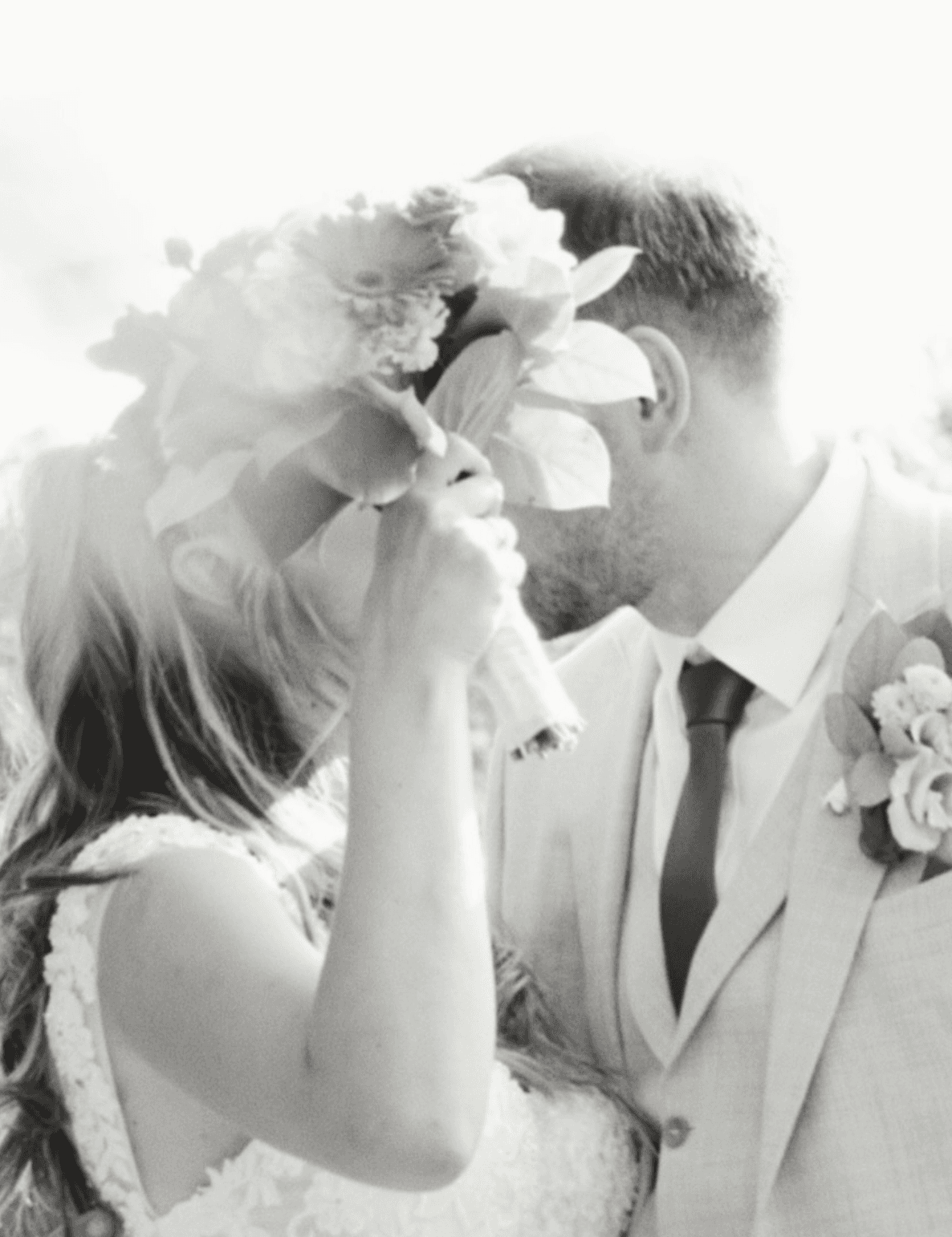 Black and white photograph of a couple embracing behind a bouquet of peonies, backlit and luminous