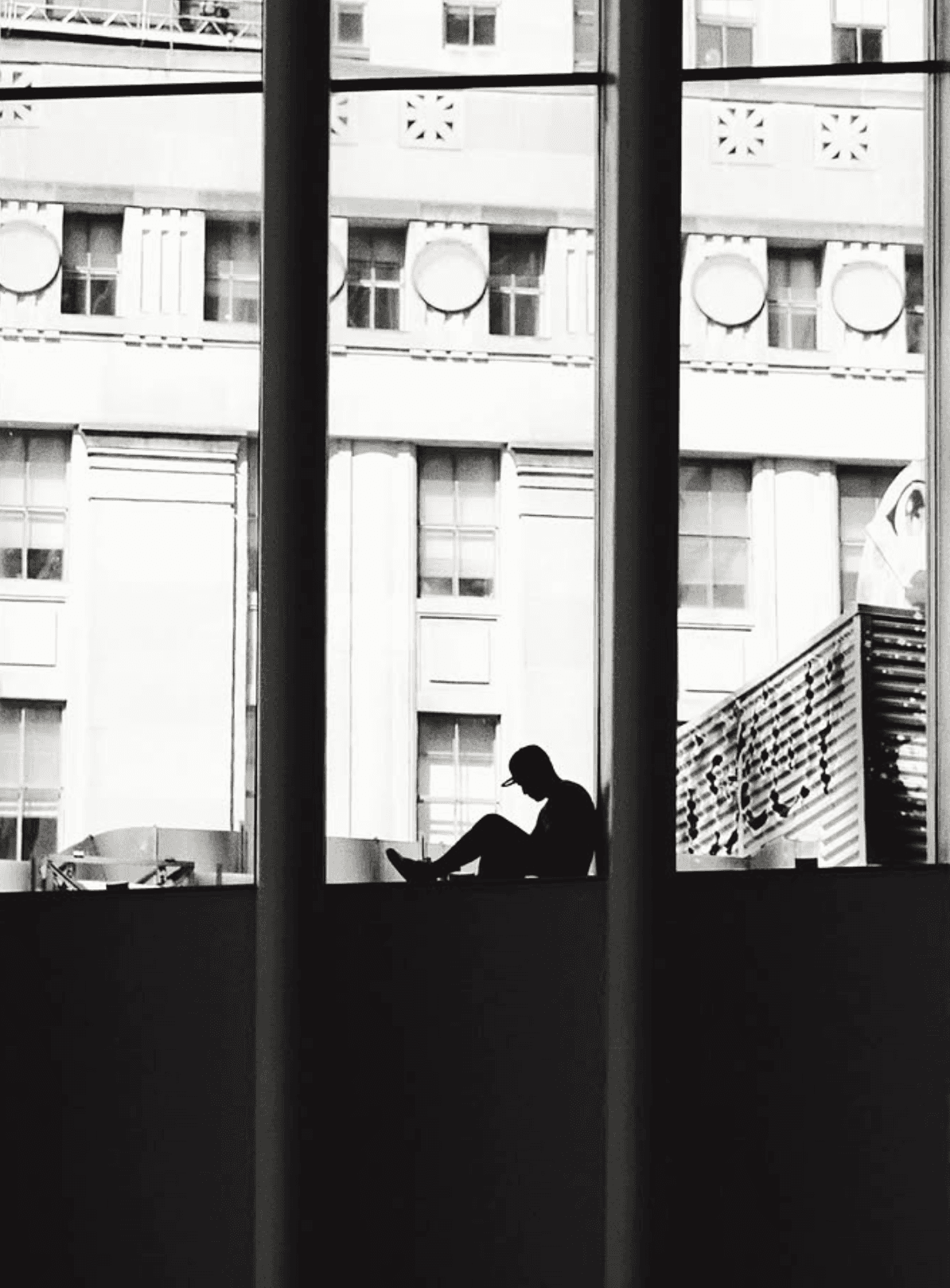 Black and white photograph of a silhouetted figure sitting in a tall city window among rows of grand facades