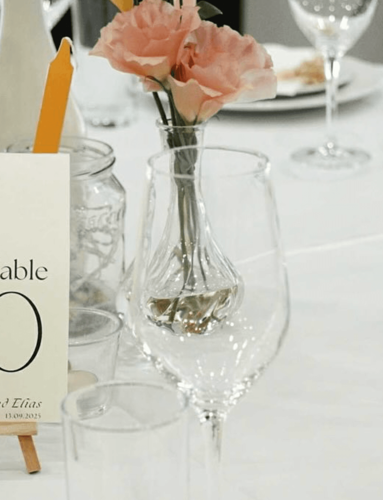 Table setting detail with wine glasses, a bud vase of pink roses and a printed table number