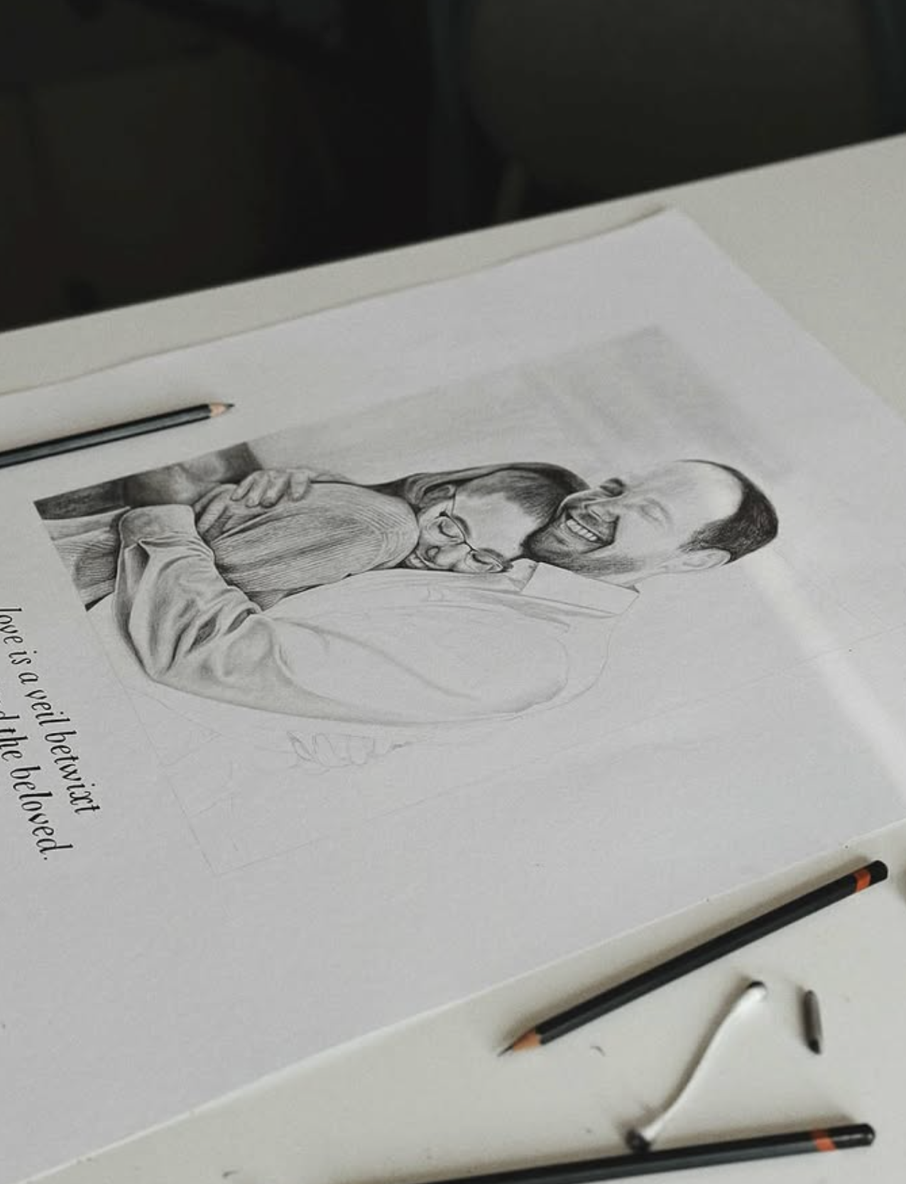 Pencil portrait of a couple mid-laugh, drawn by hand with pencils resting beside the sketchbook