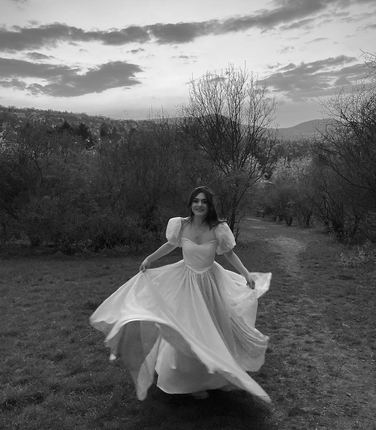 Black and white photograph of a bride twirling her dress on a hillside path at dusk