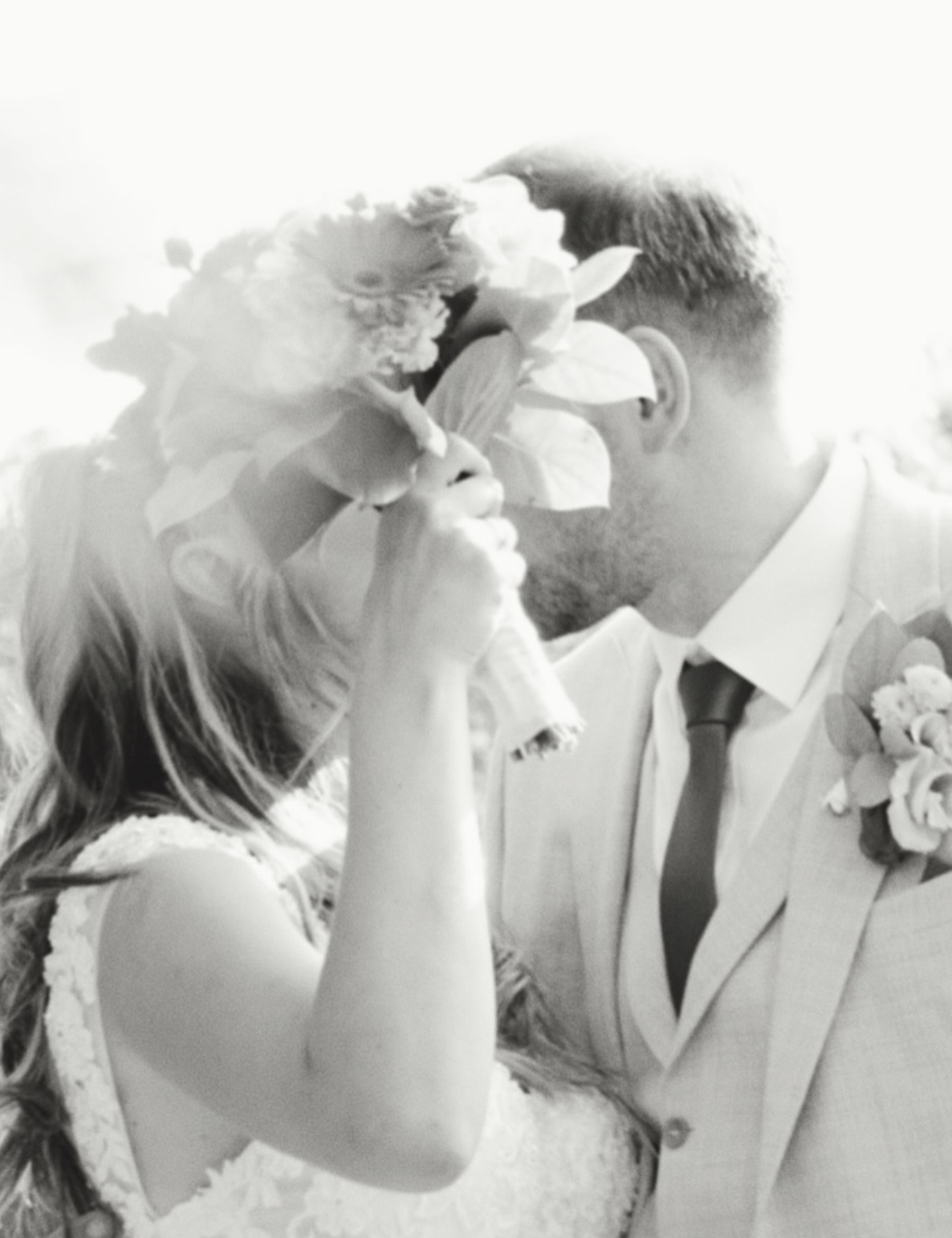 Black and white photograph of a couple embracing behind a bouquet of peonies, backlit and luminous