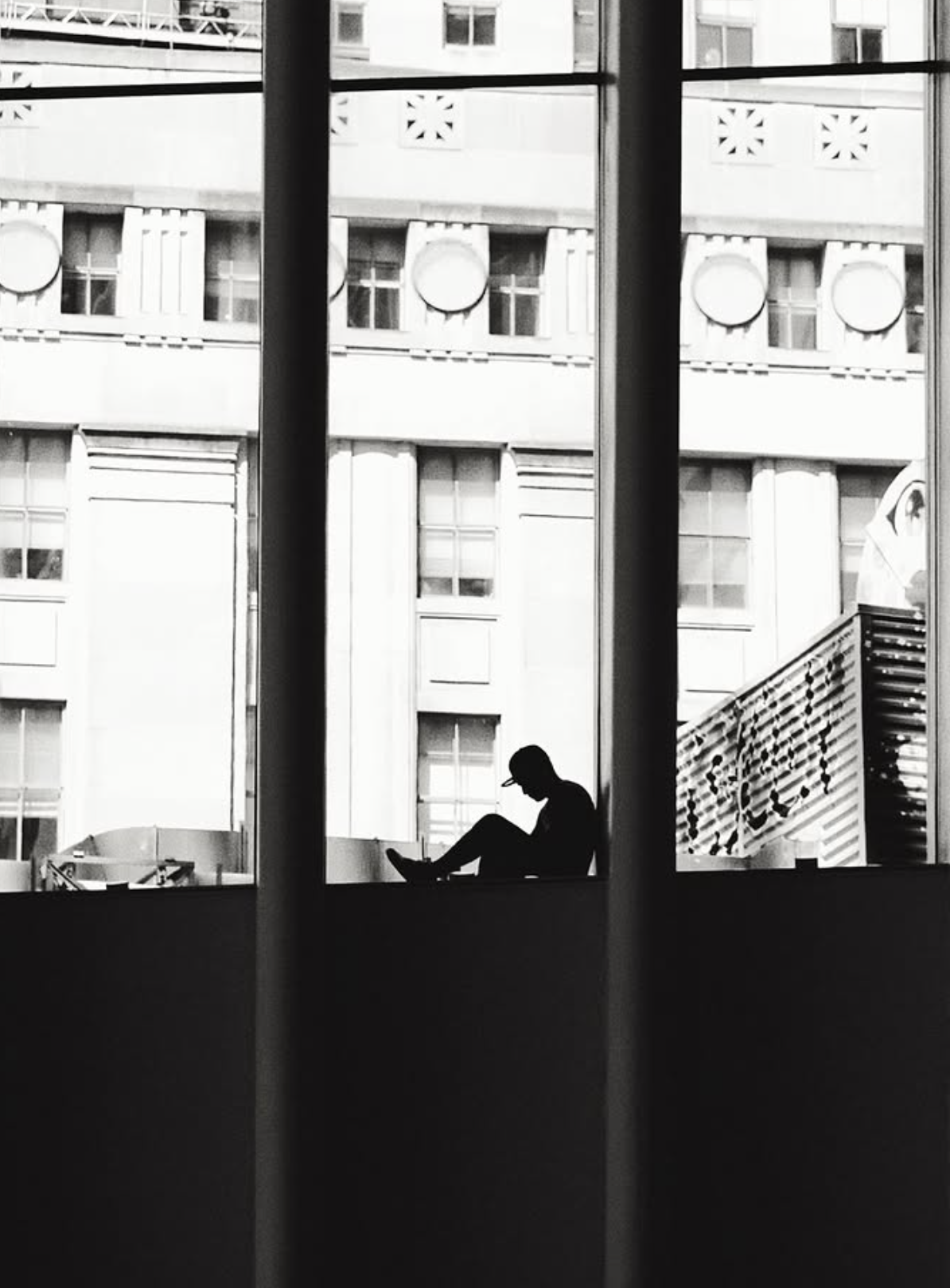 Black and white photograph of a silhouetted figure sitting in a tall city window among rows of grand facades