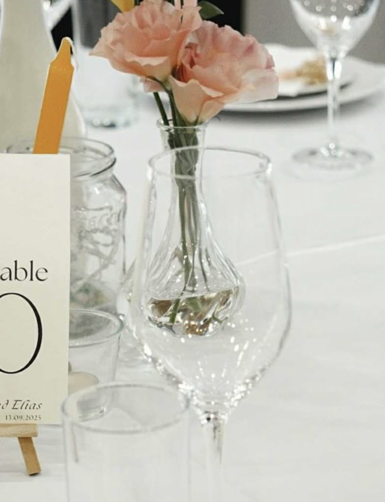 Table setting detail with wine glasses, a bud vase of pink roses and a printed table number